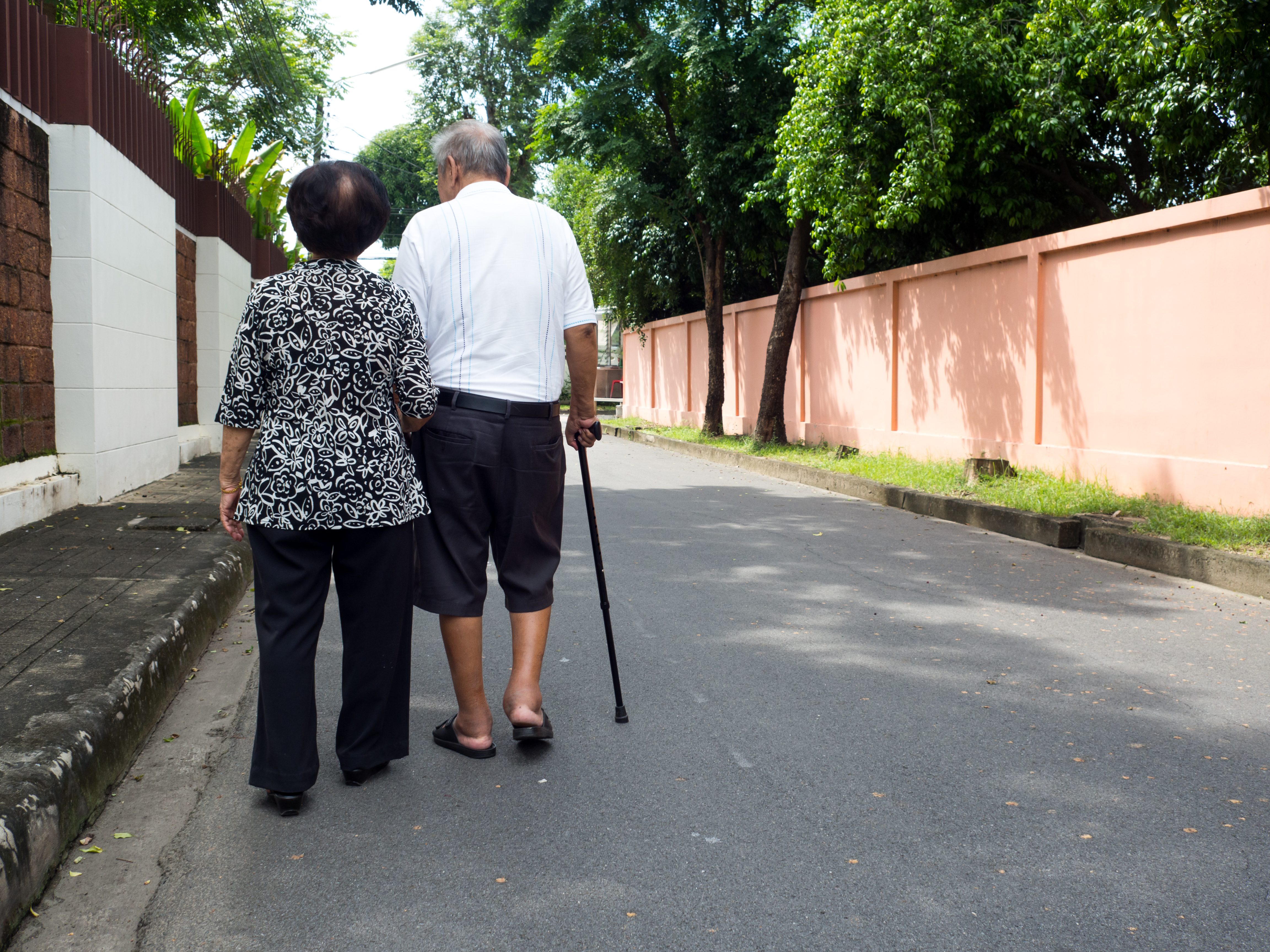 An elderly Asian couple walks down the street. The woman is holding the man's arm. The man is holding a cane with his other arm.