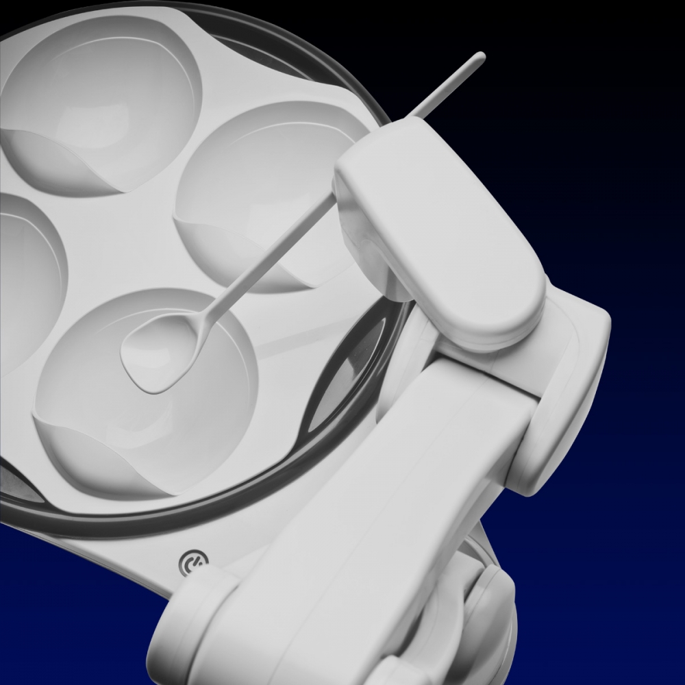 Top view of the OBI robotic arm.  It is a white mechanical arm holding a spoon, which is hovering above a plate with four sections.